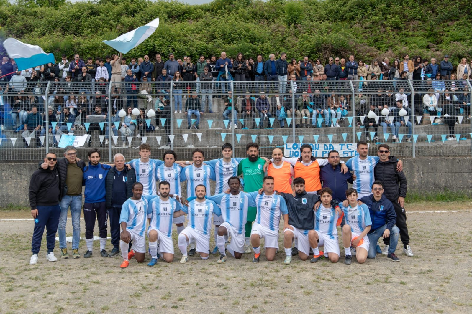 Foto di gruppo della Polisportiva Roccanova in maglia biancoceleste, con staff e dirigenti, davanti agli spalti pieni di tifosi con bandiere e festoni biancocelesti